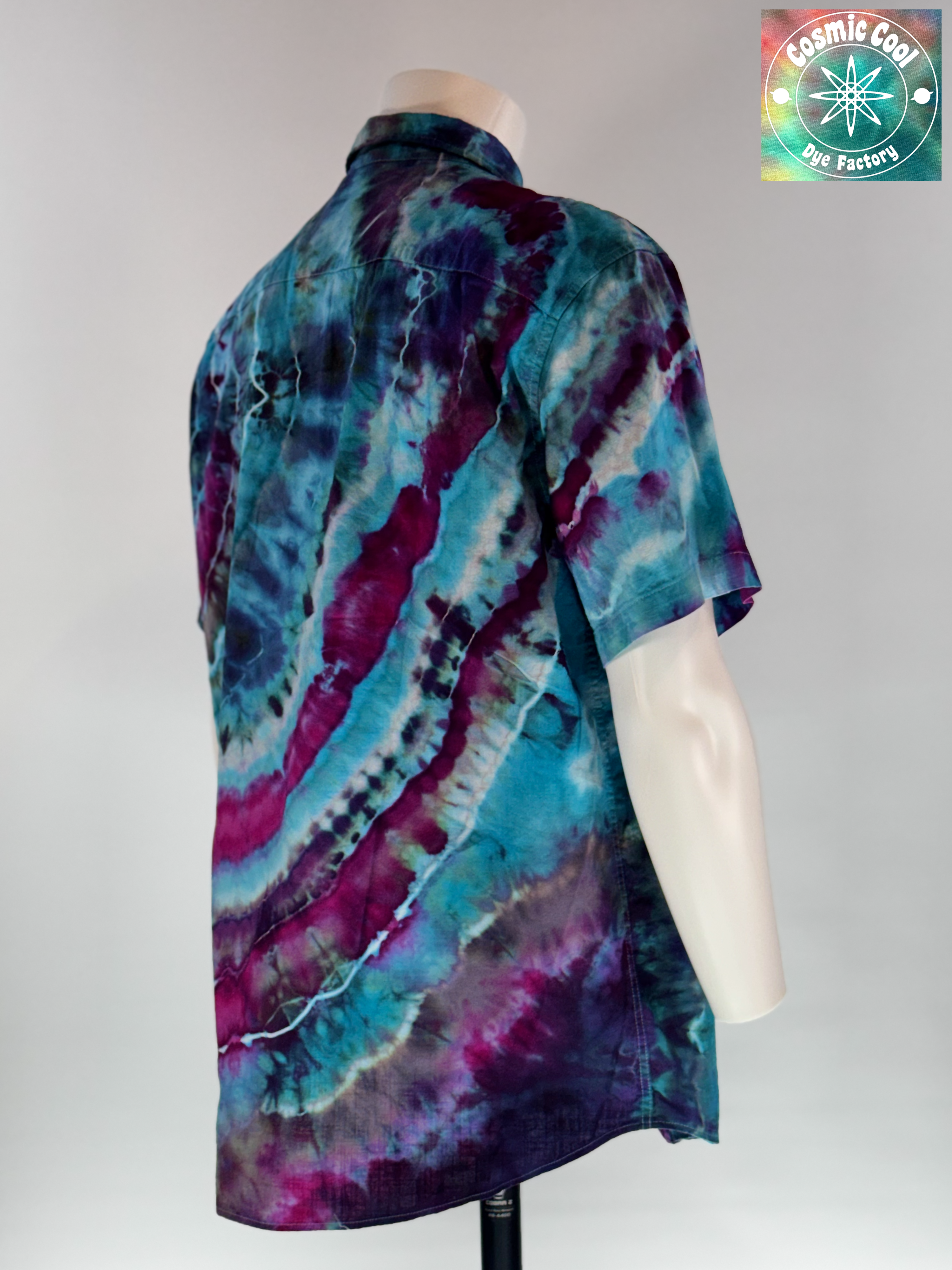 Angled 3/4 back view of our Cosmic Camp Shirt. Geodesic tides mimics the rhythmic, mineral-like layering found in coastal geodes. The design features a cascading flow of deep amethyst and royal purple waves that wash over a vibrant turquoise and teal base.
